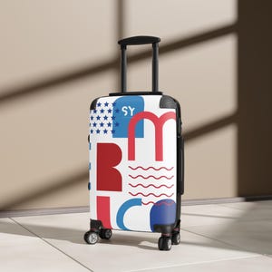Könnte beinhalten: Ein weißer Rollkoffer mit einem Design in Rot, Weiß und Blau. Das Design beinhaltet das Wort "America" in großen Buchstaben, Sterne, Streifen und die Buchstaben "SY". Der Koffer hat einen schwarzen Griff und Räder. Die Höhe beträgt ca. 55 cm.