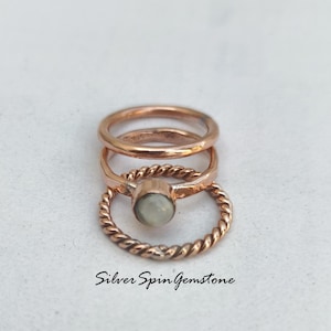 Pode incluir: Três anéis dourados com uma gema branca no centro. Os anéis estão empilhados um em cima do outro. O anel inferior é uma banda torcida. O anel do meio é uma banda lisa. O anel superior é uma banda lisa.