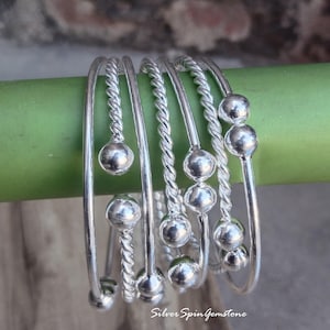 May include: A set of six silver bangles with a twisted design and small round beads. The bangles are stacked on top of each other and are arranged in a spiral pattern.