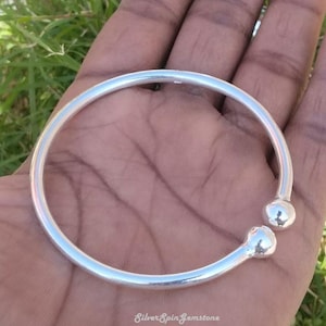 Puede incluir: Un brazalete de plata con dos bolas redondas en la abertura. El brazalete está abierto y se sostiene en una mano.