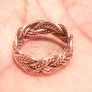 May include: A bronze-coloured ring with a braided design. The ring features a continuous pattern of interwoven strands, creating a textured and detailed appearance. The ring is a piece of jewellery.