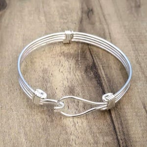 May include: A silver-colored bracelet with a hook clasp. The bracelet is made of three parallel wires, connected by wrapped sections at the ends and clasp. The bracelet is displayed on a wooden surface.