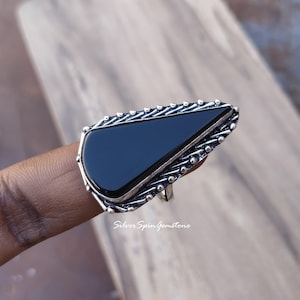May include: A silver ring with a large, triangular black onyx stone. The ring has a detailed, textured border around the stone. The ring is displayed on a finger. The text "Silver Spin Gemstone" is visible on the image.