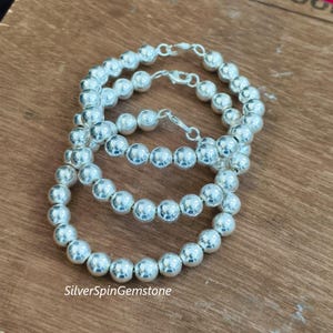 May include: Three silver beaded bracelets with a polished finish are displayed on a wooden surface. Each bracelet is composed of round, evenly sized beads and features a clasp closure. The text "SilverSpinGemstone" is visible.