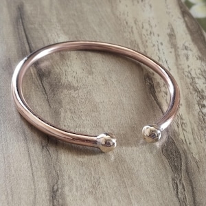 Könnte beinhalten: Ein schlichtes Roségold-Armreif-Armband mit abgerundeten Enden. Das Armband besteht aus glattem, poliertem Metall.