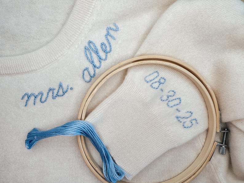 May include: A white knitted sweater with blue embroidery that reads "mrs. allen" and "08-30-25". The sweater is being held in place by a wooden embroidery hoop.