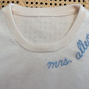 May include: A white knitted sweater with blue embroidery that reads "mrs. allen".