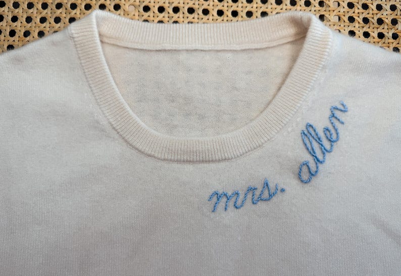 May include: A white knitted sweater with blue embroidery that reads "mrs. allen".