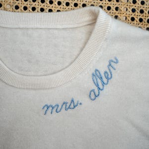 May include: A white knitted sweater with blue embroidery that reads "mrs. allen".