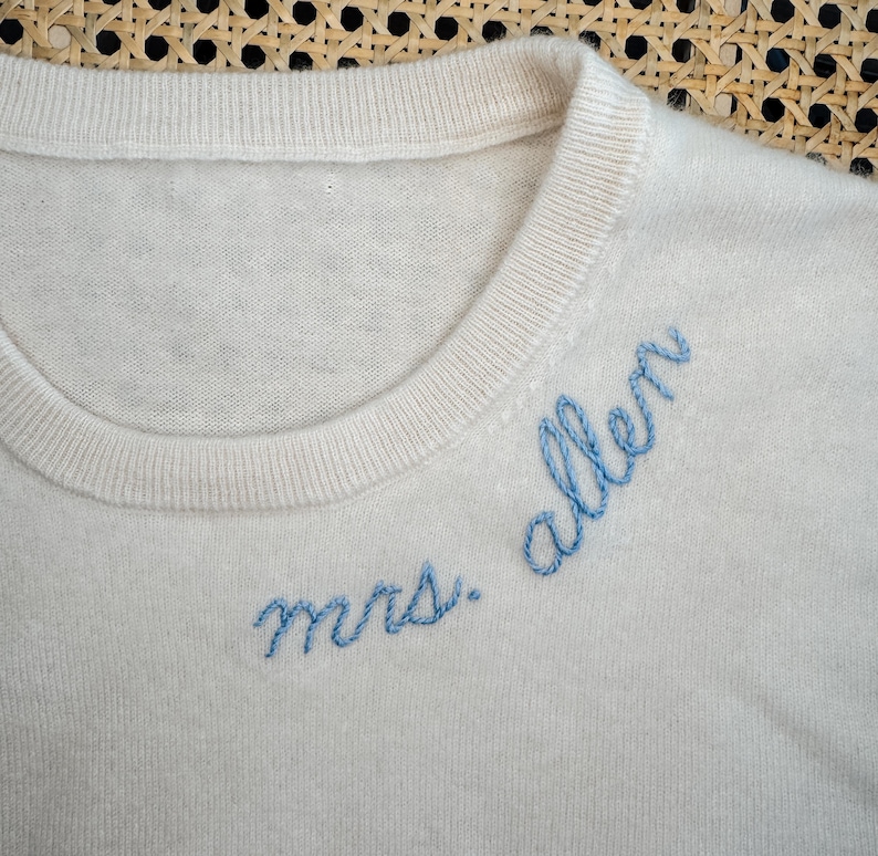 May include: A white knitted sweater with blue embroidery that reads "mrs. allen".