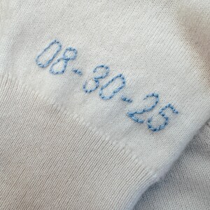 May include: White knitted fabric with blue embroidered text that reads "08.30.23".