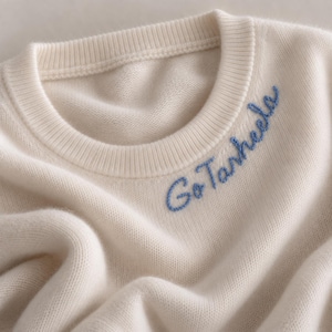 May include: A cream-coloured crew neck jumper featuring the text Go Tarheels embroidered in blue thread on the upper right chest area. The garment has a ribbed neckline and a soft, knit texture throughout the fabric surface.
