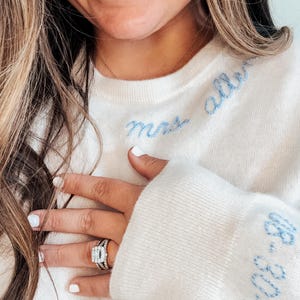 May include: A white sweater with blue embroidery that says "Mrs. Allen" on the front and "Est. 2023" on the sleeve. The sweater is worn by a person with a ring on their finger.