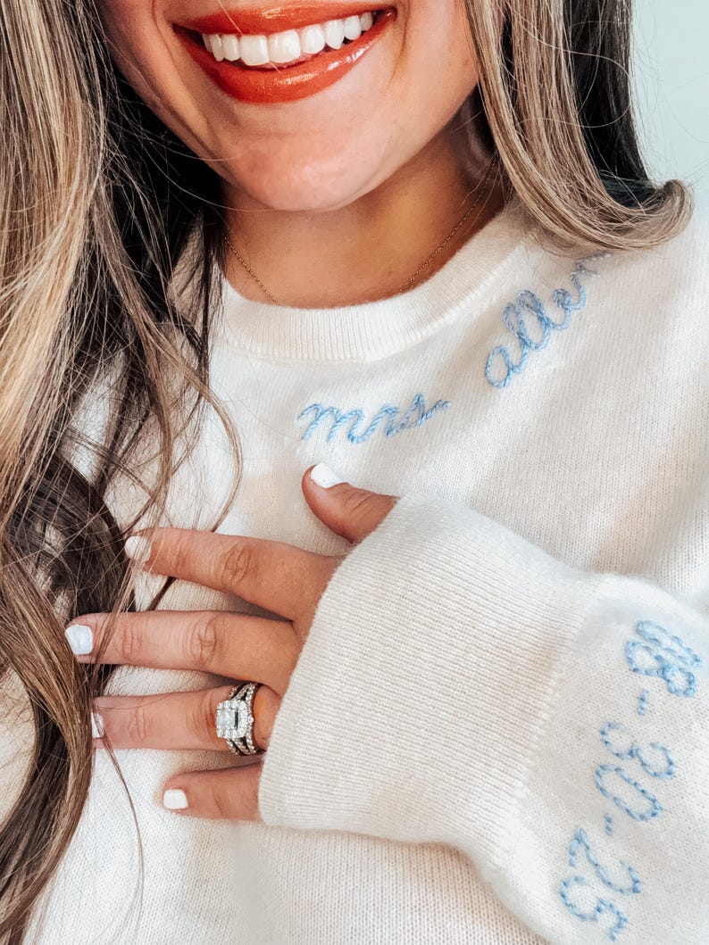 May include: A white sweater with blue embroidery that says "Mrs. Allen" on the front and "Est. 2023" on the sleeve. The sweater is worn by a person with a ring on their finger.