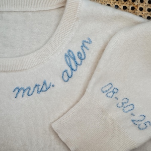 May include: A white knitted sweater with blue embroidery that reads "mrs. allen" and "08-30-23".