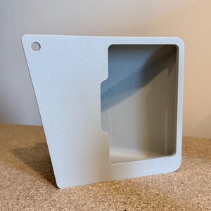 May include: A light gray, angular, open-faced container with a rectangular opening and a small hole in the upper left corner. The container has a smooth, matte finish and appears to be made of a solid material.