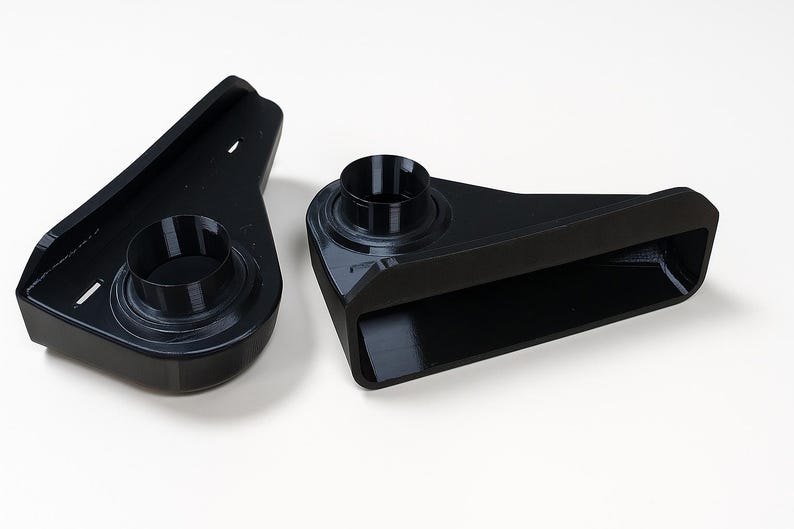 Puede incluir: Dos componentes de pl&aacute;stico negro, angulares, con aberturas cil&iacute;ndricas. Una pieza tiene una extensi&oacute;n rectangular. Los objetos est&aacute;n sobre una superficie blanca. El dise&ntilde;o sugiere una aplicaci&oacute;n mec&aacute;nica o t&eacute;cnica.