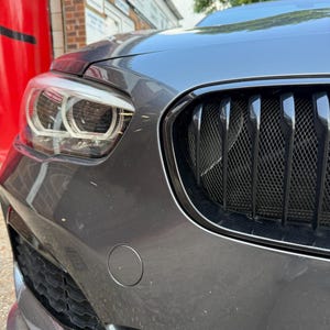 May include: Close-up of a gray car's front end, showcasing the headlight, black grill, and bumper. The car's body has a metallic finish. The image is taken outdoors on a sunny day.
