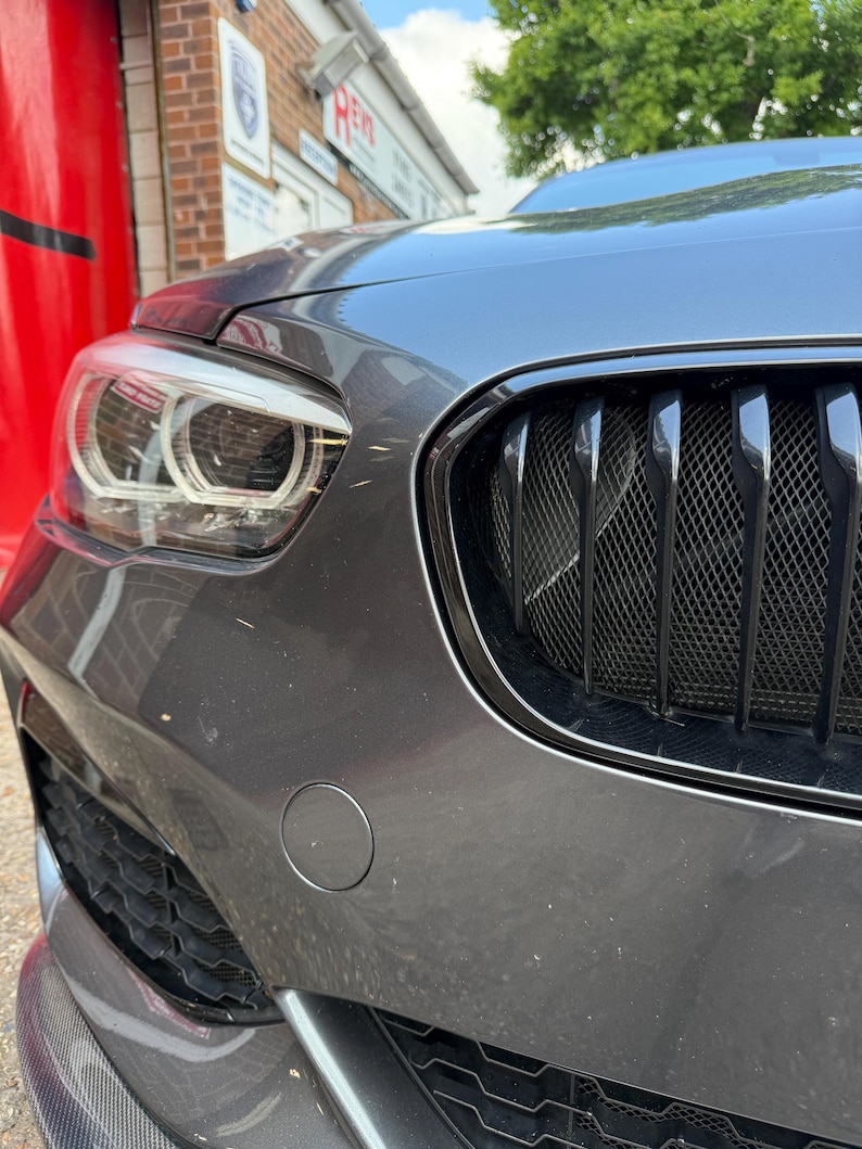 May include: Close-up of a gray car's front end, showcasing the headlight, black grill, and bumper. The car's body has a metallic finish. The image is taken outdoors on a sunny day.