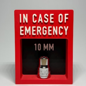 Puede incluir: Una pantalla rectangular roja con las palabras "IN CASE OF EMERGENCY" en letras blancas en relieve. En el interior, un fondo negro muestra "10 MM" y una pequeña herramienta plateada y roja.