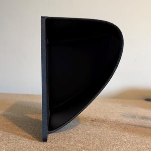 May include: A black, curved, wall-mounted shelf with a textured surface. The shelf has a rounded front edge and a flat back, designed to hold small items. The shelf is made of a solid material.