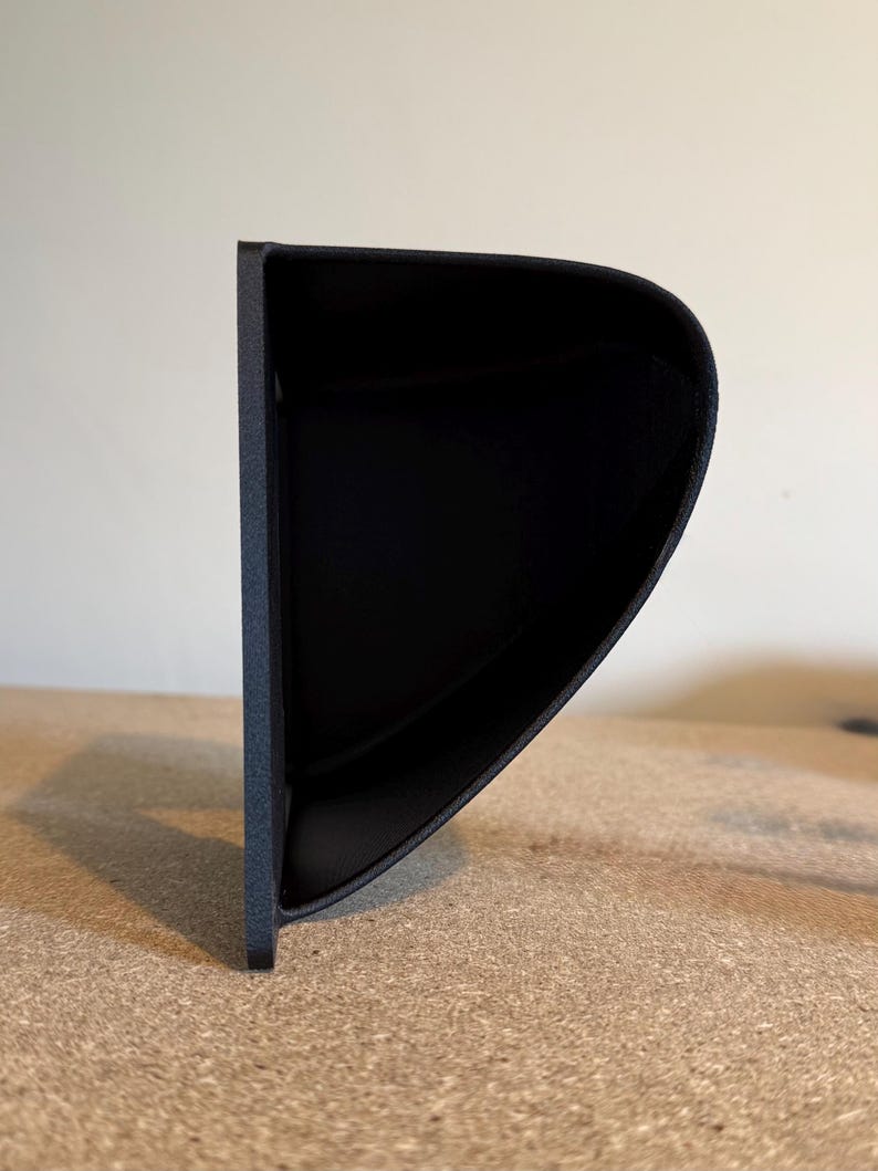 May include: A black, curved, wall-mounted shelf with a textured surface. The shelf has a rounded front edge and a flat back, designed to hold small items. The shelf is made of a solid material.
