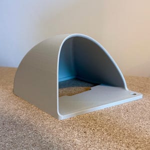 May include: A light gray, semi-circular shelf with a flat base and a small hole on the right side. The shelf has a smooth, matte finish and is made of a solid material. The shelf is sitting on a textured, brown surface.