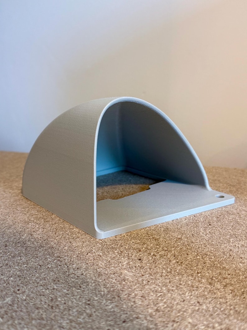 May include: A light gray, semi-circular shelf with a flat base and a small hole on the right side. The shelf has a smooth, matte finish and is made of a solid material. The shelf is sitting on a textured, brown surface.
