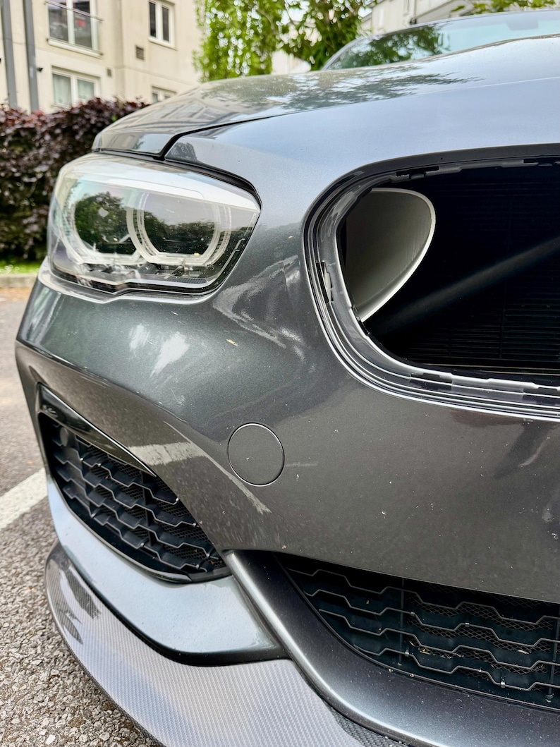 May include: Close-up of a gray car's front end, showcasing the headlight, grill, and bumper. The headlight is illuminated, and the grill has a honeycomb pattern. The bumper has a carbon fiber finish, and the car appears to be parked outdoors.