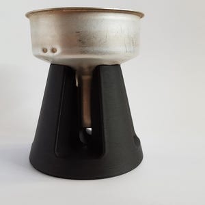 Op de afbeelding: Een metalen koffiefilter staat op een zwarte, kegelvormige standaard. De filter is zilverkleurig met een afgeronde rand en twee kleine gaatjes. De standaard heeft een gestructureerd oppervlak en een brede basis.