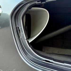 May include: A close-up view of a car's front end, showcasing a white, curved plastic component within the vehicle's frame. The car's body is a metallic gray, and the image highlights the integration of the part.