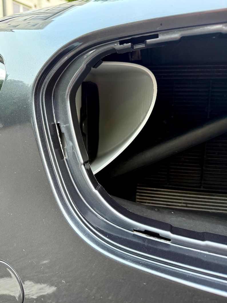 May include: A close-up view of a car's front end, showcasing a white, curved plastic component within the vehicle's frame. The car's body is a metallic gray, and the image highlights the integration of the part.