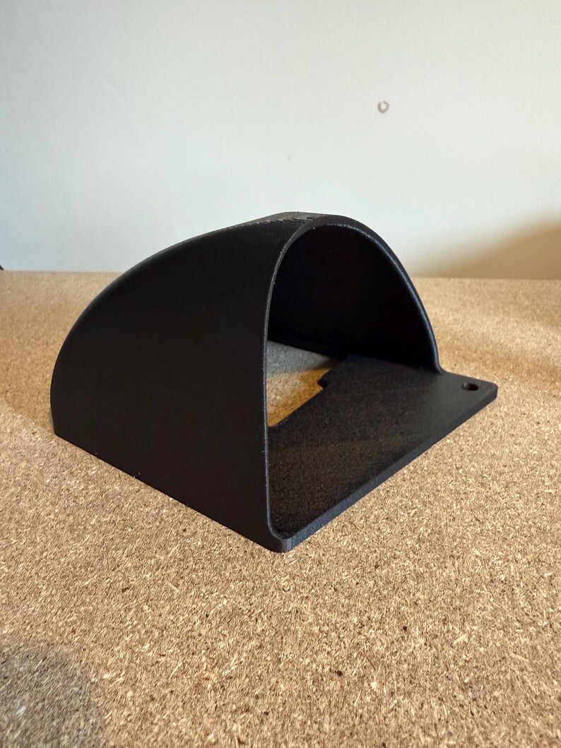 May include: A black, curved metal bracket with a flat base and a single mounting hole. The bracket has a rounded top and a rectangular opening, suitable for mounting or holding an item. The surface has a textured finish.