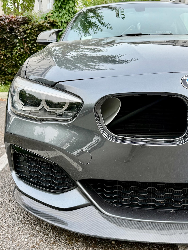 May include: Close-up of a gray BMW car's front end. The car features a black grill, a carbon fiber front lip, and a sleek headlight design. The car is parked on a paved surface, with greenery visible in the background.