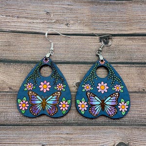 May include: A pair of blue wooden earrings shaped like a witch's board or planchette. Each earring features a butterfly with pink, blue, and purple wings, surrounded by pink and yellow flowers and green leaves.