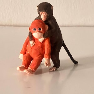 May include: Two small monkey figurines. One is a vibrant orange, the other is dark brown. The brown monkey is embracing the orange monkey from behind. The figurines are on a white surface.