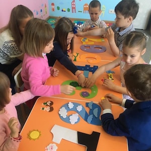 Puede incluir: Niños ensamblando números de fieltro de colores en una mesa naranja. Los números son de varios colores, incluyendo verde, azul y amarillo. Los niños están concentrados en la actividad, algunos sosteniendo piezas.