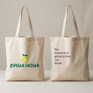 May include: Two off-white canvas tote bags with two shoulder straps each. The left bag features a yellow pear graphic and the green text "NASHI HOSHI". The right bag displays the purple text "Not everyone is going to love you. Good."