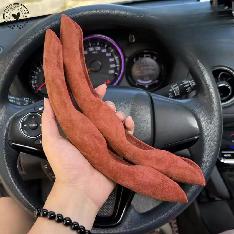 May include: Two rust-colored, curved car accessories are held in front of a car's steering wheel. The accessories have a soft, velvety texture. The car's dashboard and steering wheel are visible in the background.