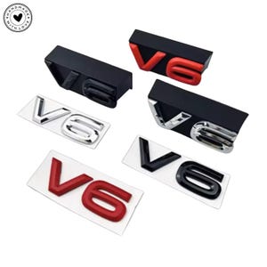 Puede incluir: Una colección de emblemas de coche V6 en varios estilos. Algunos son negros con letras plateadas, otros negros con letras rojas y algunos plateados con letras negras. Todos sobre fondo blanco.