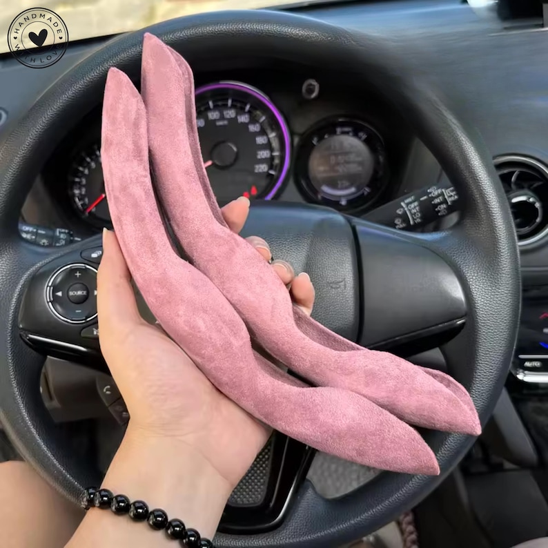 May include: Two pink suede steering wheel covers are held in front of a car's dashboard. The covers are curved and shaped to fit the steering wheel. The car's speedometer and other gauges are visible in the background.