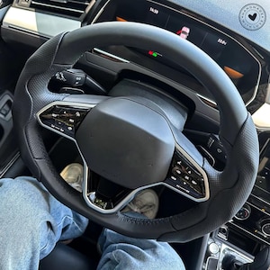 May include: Black leather steering wheel with silver accents and integrated controls. The wheel has a perforated grip section and a central airbag cover. The dashboard displays a digital screen with vehicle information.
