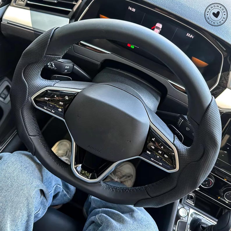 May include: Black leather steering wheel with silver accents and integrated controls. The wheel has a perforated grip section and a central airbag cover. The dashboard displays a digital screen with vehicle information.