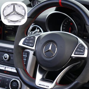 Può includere: Un volante in pelle nera con un logo Mercedes-Benz argentato e cuciture rosse. Il volante ha i comandi per il sistema audio dell'auto e il cruise control. Il volante ha anche la scritta "AMG" stampata su di esso.