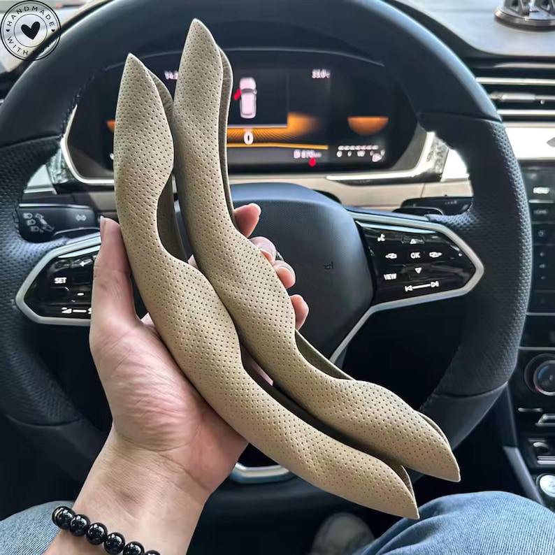 May include: Two tan, perforated leather steering wheel covers. The covers are held in front of a car's steering wheel, showcasing their curved shape and textured surface. The covers are designed to fit the steering wheel.