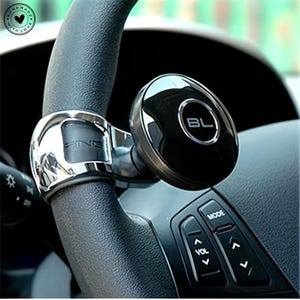 May include: A black and chrome steering wheel spinner attached to a black steering wheel. The spinner has the letters "BL" in a silver circle. The steering wheel has buttons for mode, volume, and directional controls.
