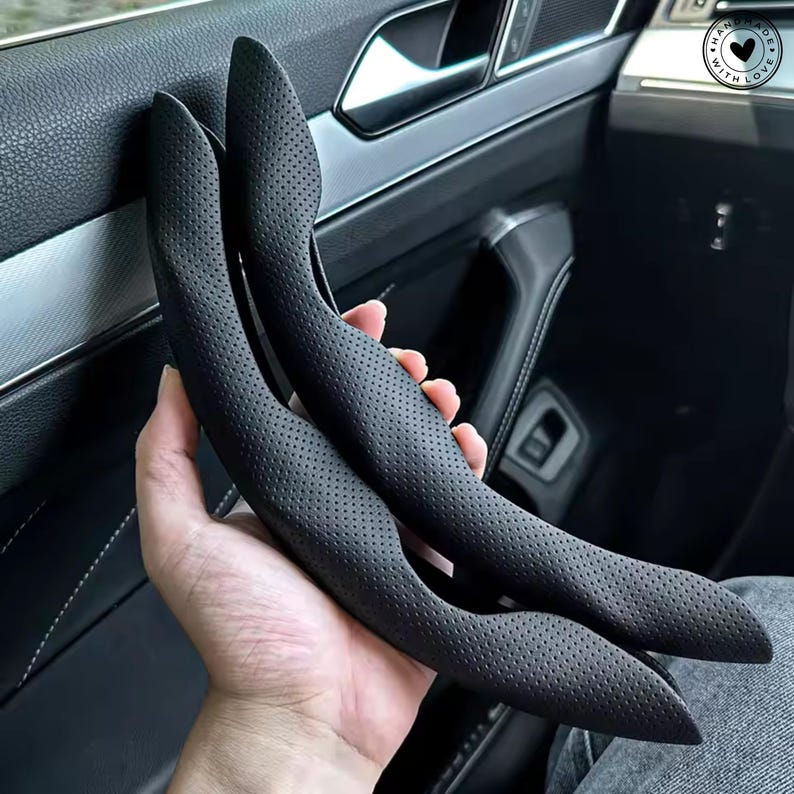 May include: Two black, perforated leather car door handle covers. The covers are curved and designed to fit over the existing handles. The image shows the covers held in a hand, with the interior of a car in the background.
