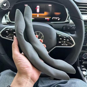 May include: Two gray, perforated leather steering wheel covers. The covers are curved and designed to fit the steering wheel. The background shows the car's dashboard and steering wheel.