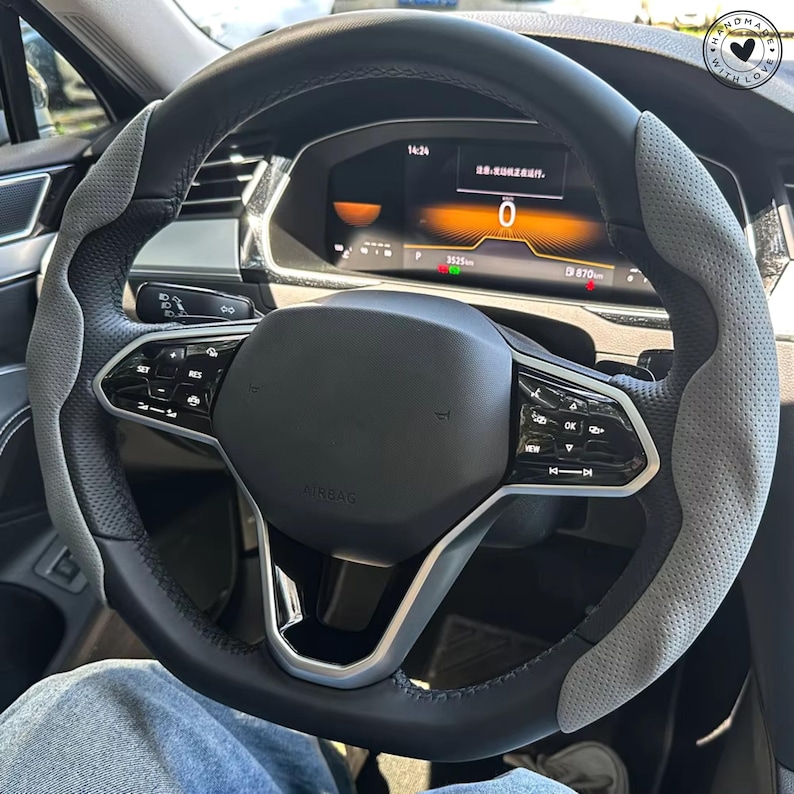 May include: A black and gray steering wheel with a perforated gray grip section. The wheel has silver accents and is set against a car dashboard displaying digital information. The airbag cover is black, and the wheel has various control buttons.