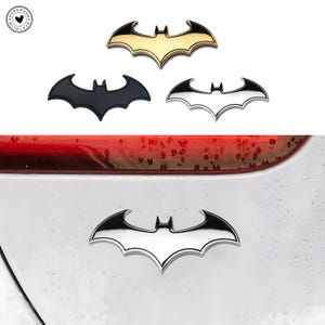 May include: Four Batman logo emblems in gold, black, and silver. The emblems are in the shape of a bat with outstretched wings. One emblem is attached to a white surface.